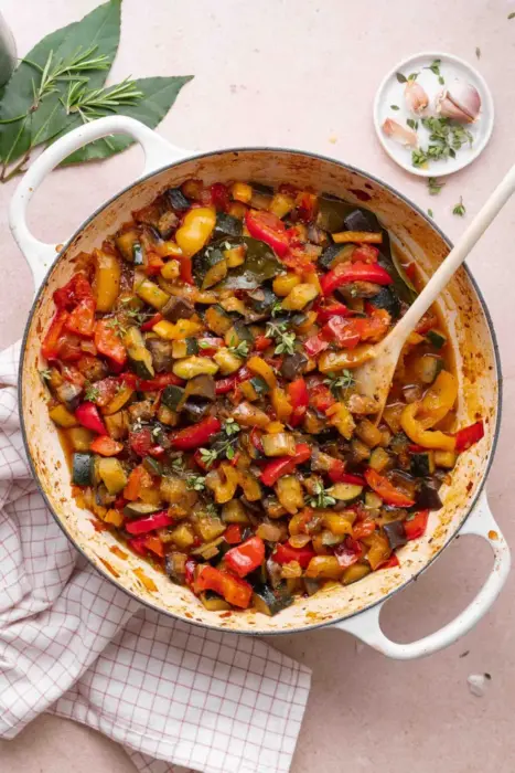 Ratatouille i en vit stor gryta. På bilden syns även en kökshandduk, vitlök upplagt bredvid samt några gröna blad.